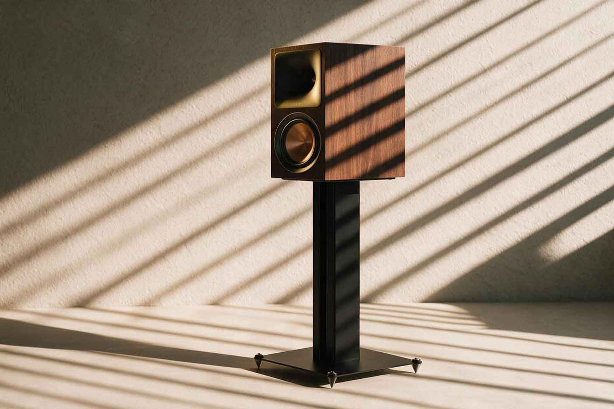 One  wood-finished tower loudspeaker with flush horn tweeters and metal outrigger feet, lit at an angle with long shadows.